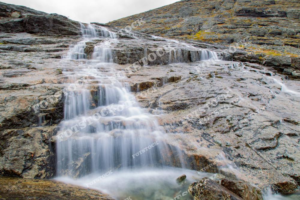 На фотографии изображен горный водопад, который спускается по каменистым склонам Хибин. Вода плавно стекает, создавая живописные потоки среди камней, что является характерным для северных горных регионов России.